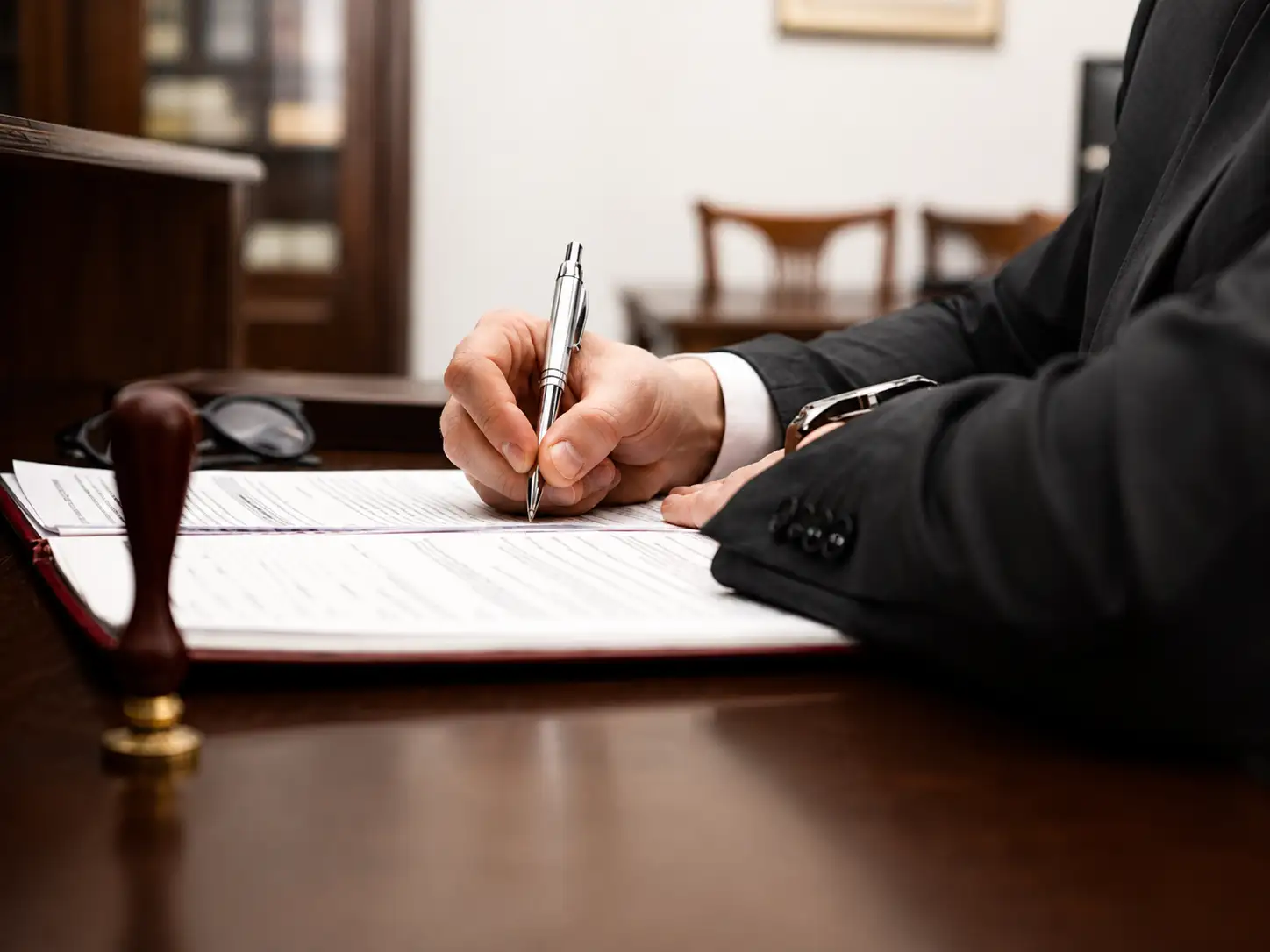 Person in a dark suit signs a document with a silver pen at a wooden desk, a stamp visible in the foreground.