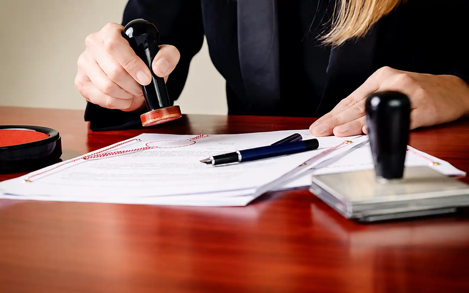 Person stamps a stack of official papers on a wooden desk, with a pen nearby and another stamp visible on the side.