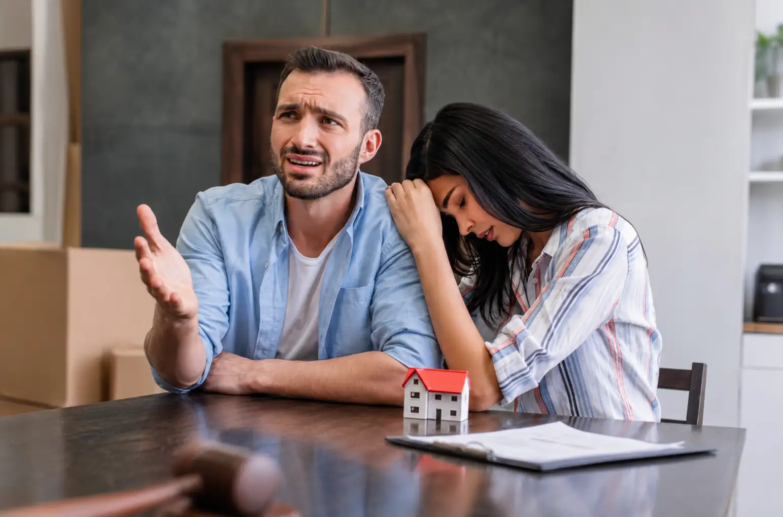 Couple arguing at a kitchen/dining table with a small house model, documents, and a gavel, suggesting a real estate dispute or decision.